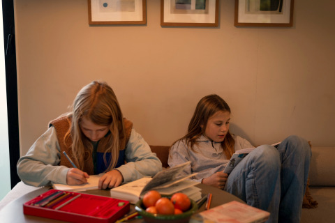 Deux enfants se détendent dans un hébergement glamping, l’un écrit et l’autre lit, avec livres et fruits.