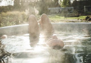Une personne se détend dans un bain chaud fumant en plein air, entourée de verdure dans un lodge.