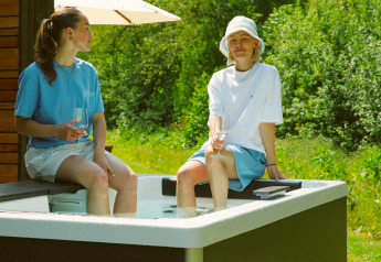 Zwei Personen sitzen am Rand eines Außenwhirlpools an einer Lodge und genießen Getränke bei Sonnenschein.
