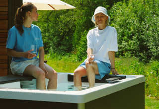 Deux personnes assises au bord d’un jacuzzi extérieur à une cabane, dégustant un verre sous le soleil.