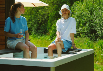 To personer sidder på kanten af et udendørs badekar ved en lodge og nyder drikkevarer en solrig dag.