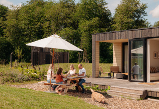 Gezin eet buiten aan een picknicktafel bij een lodge, genietend van het zonnetje en een spelende hond.