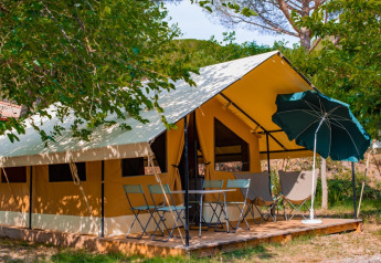 Tenda safari Ponza con terrazza esterna, tavolo, sedie e ombrellone sotto alberi frondosi.