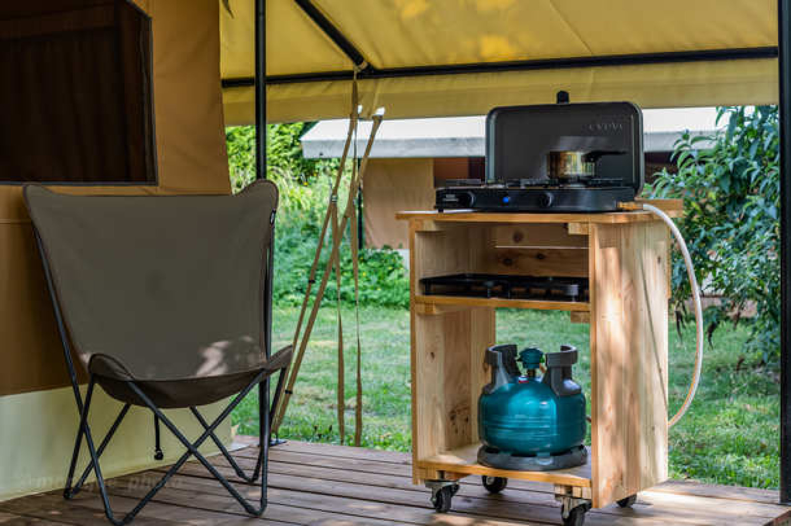 Glampingplek met campingstoel en gasfornuis op houten kar, omringd door groen en natuur.