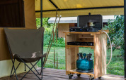 Udendørs glamping område med campingstol og gaskomfur på trækøkkenvogn, omgivet af grøn natur.