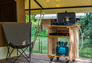 Coin glamping avec fauteuil de camping et réchaud à gaz sur chariot en bois, dans un cadre verdoyant.
