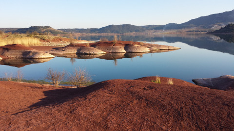 Red earth hills and calm water with distant mountains in Lodève, Occitanie, France, on a clear day.