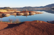 Colinas de tierra roja junto a aguas tranquilas con montañas al fondo cerca de Lodève, Occitania, Francia.