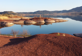 Red earth hills and calm water with distant mountains in Lodève, Occitanie, France, on a clear day.