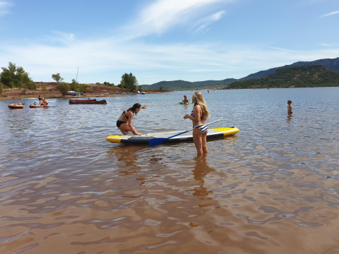 Dos mujeres hacen paddle surf en aguas poco profundas, con personas y barcos al fondo en un lago de Occitanie, Francia.
