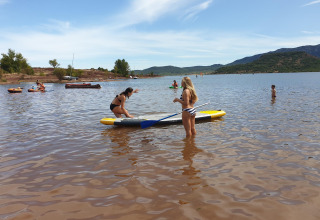 Twee vrouwen op een paddleboard in ondiep water, met zwemmers en boten op een meer in Occitanië, Frankrijk.