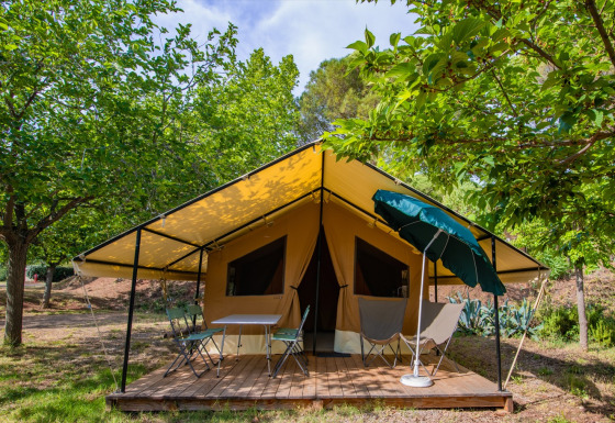 A Ponza safari tent with yellow canopy, terrace, chairs, and umbrella surrounded by lush trees.