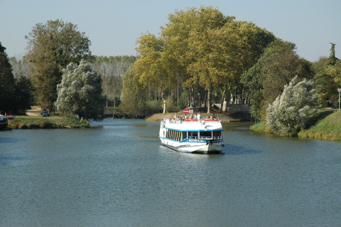 A tourist boat cruises a calm river bordered by trees near Onlycamp Camping le Champ d'Eté holiday park, France.