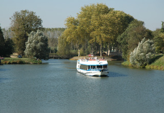Een rondvaartboot vaart op een rustige rivier omringd door bomen bij Onlycamp Camping le Champ d'Eté in Frankrijk.