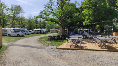 Outdoor campsite with caravans, cars and shaded seating areas under trees on a sunny day.