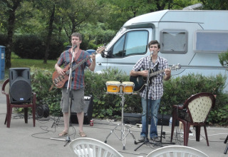 Dos músicos tocan guitarras y cantan con micrófonos al aire libre frente a una caravana en Onlycamp Camping de Besançon.