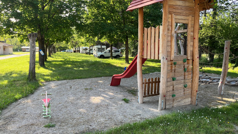 Playground with wooden playhouse, slide, and swings under shady trees at Onlycamp Camping Chamarges, France.