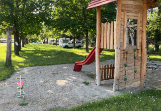 Playground with wooden playhouse, slide, and swings under shady trees at Onlycamp Camping Chamarges, France.