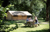 Familie sitzt am Picknicktisch vor einem Ponza-Safari-Zelt und genießt die Natur auf einem grünen Campingplatz.