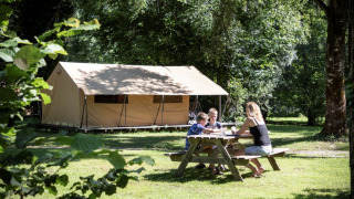 Familie sitzt am Picknicktisch vor einem Ponza-Safari-Zelt und genießt die Natur auf einem grünen Campingplatz.