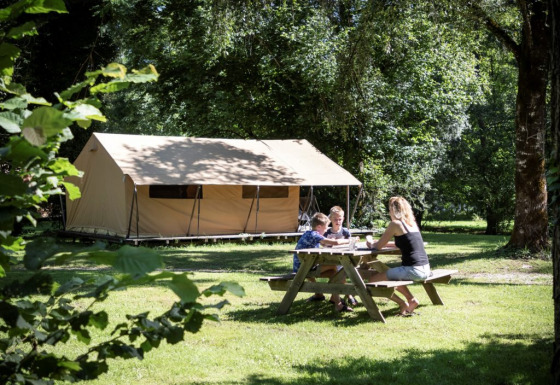 Familie sitzt am Picknicktisch vor einem Ponza-Safari-Zelt und genießt die Natur auf einem grünen Campingplatz.