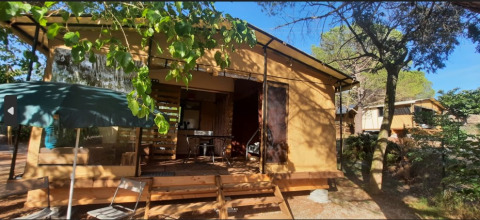 Tienda tipo safari con terraza de madera, sillas y sombrilla al aire libre en entorno boscoso soleado.