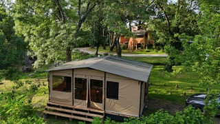 Tenda safari su piattaforma in legno tra alberi verdi, con una casa sull'albero sullo sfondo visibile.