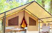Glampingverblijf in het bos met een grote tent, houten terras, stoelen en tafel onder de bomen.