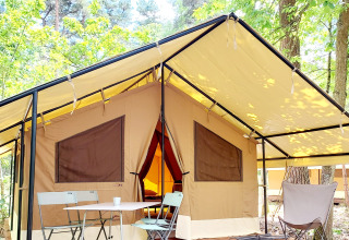 Skøn glamping-indkvartering i skoven med stort telt, veranda, bord og stole under trækronerne.