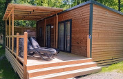 A modern wooden lodge with a covered terrace and loungers, surrounded by trees in a peaceful forest setting.