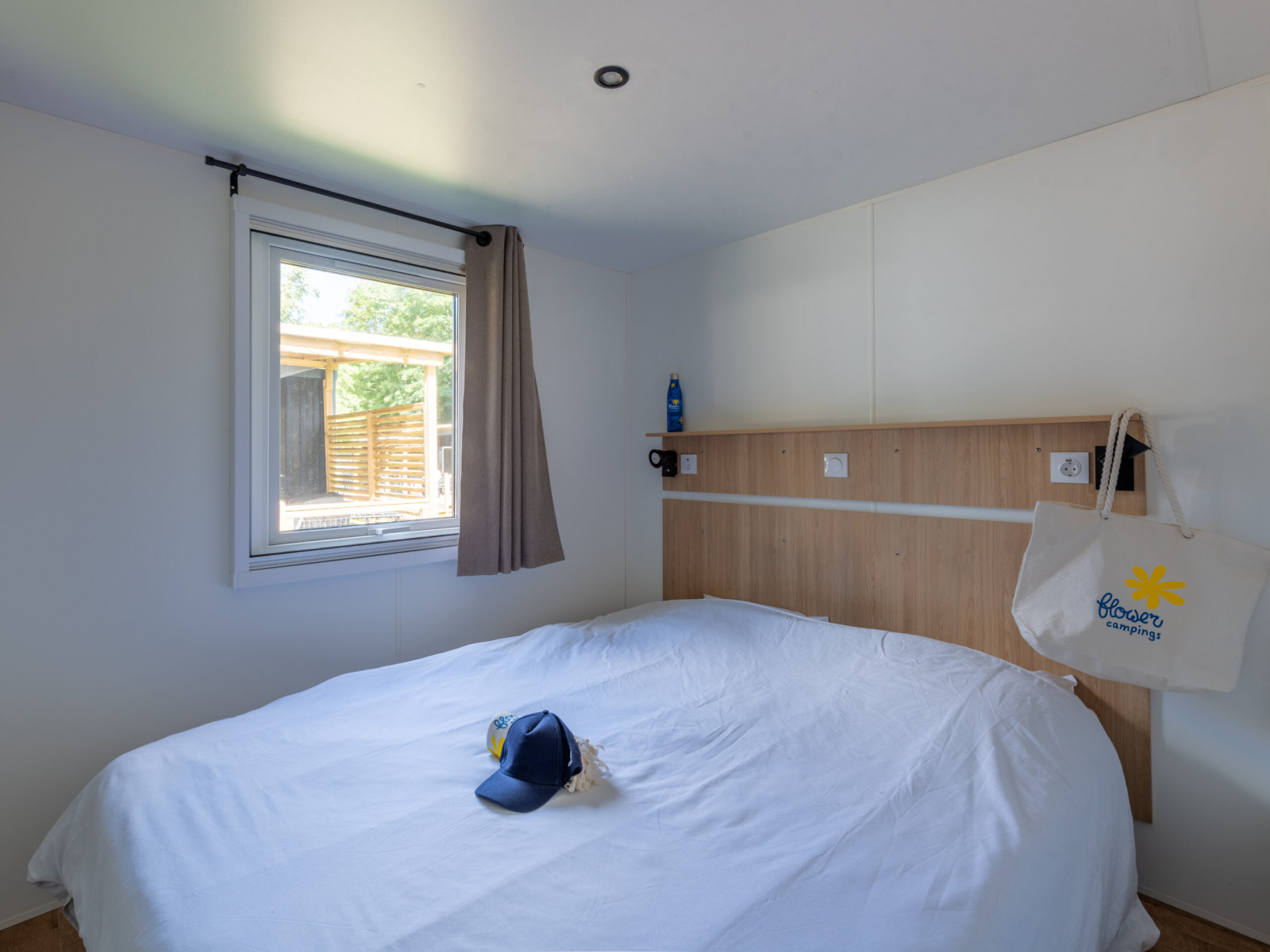 Lichte lodgekamer met tweepersoonsbed, wit beddengoed, raam en een Flower Camping tas aan het hoofdbord.