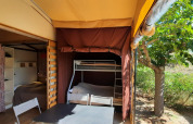 Hébergement glamping avec lit superposé, table et chaises sous auvent, vue sur arbre et verdure.