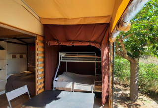 Glamping indkvartering med køjeseng, bord og stole under teltdug, udendørs træ og naturlig udsigt.
