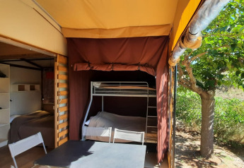 Glamping indkvartering med køjeseng, bord og stole under teltdug, udendørs træ og naturlig udsigt.