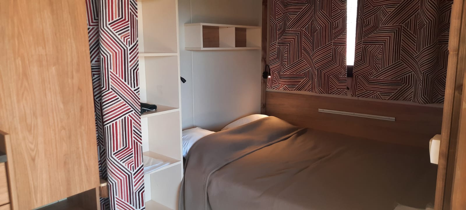 Knusse glampingaccommodatie met tweepersoonsbed, open planken en gordijnen met geometrisch patroon.