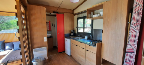 Moderne Glamping-Küche mit Holzoptik, Gasherd, kleinem Kühlschrank und großen Fenstern mit Tageslicht.