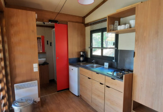Moderne Glamping-Küche mit Holzoptik, Gasherd, kleinem Kühlschrank und großen Fenstern mit Tageslicht.