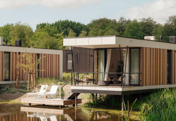 Moderne glampingaccommodatie met terras boven het water, omringd door natuur en ligstoelen buiten.
