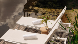 Deux chaises longues avec serviettes blanches au bord d’un étang dans un hébergement glamping, vase fleuri.