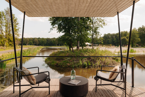 Blick von einer Terrasse mit zwei Stühlen am Wasser bei einem Glampingplatz, umgeben von Natur.