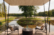Blick von einer Terrasse mit zwei Stühlen am Wasser bei einem Glampingplatz, umgeben von Natur.