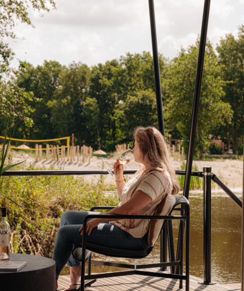 Frau entspannt mit einem Glas Wein auf der Terrasse einer Glamping-Unterkunft am Wasser.