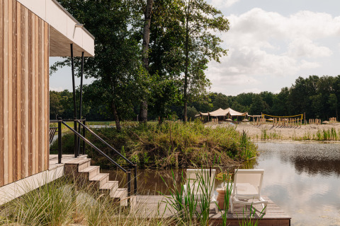 Glamping-Unterkunft am See mit Holzterrasse, Liegestühlen und Sandstrand, umgeben von Bäumen.