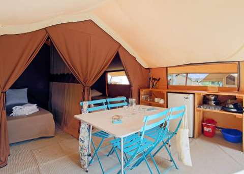 Intérieur de la tente safari Ponza avec table à manger, chaises bleues, lits et coin cuisine compact.