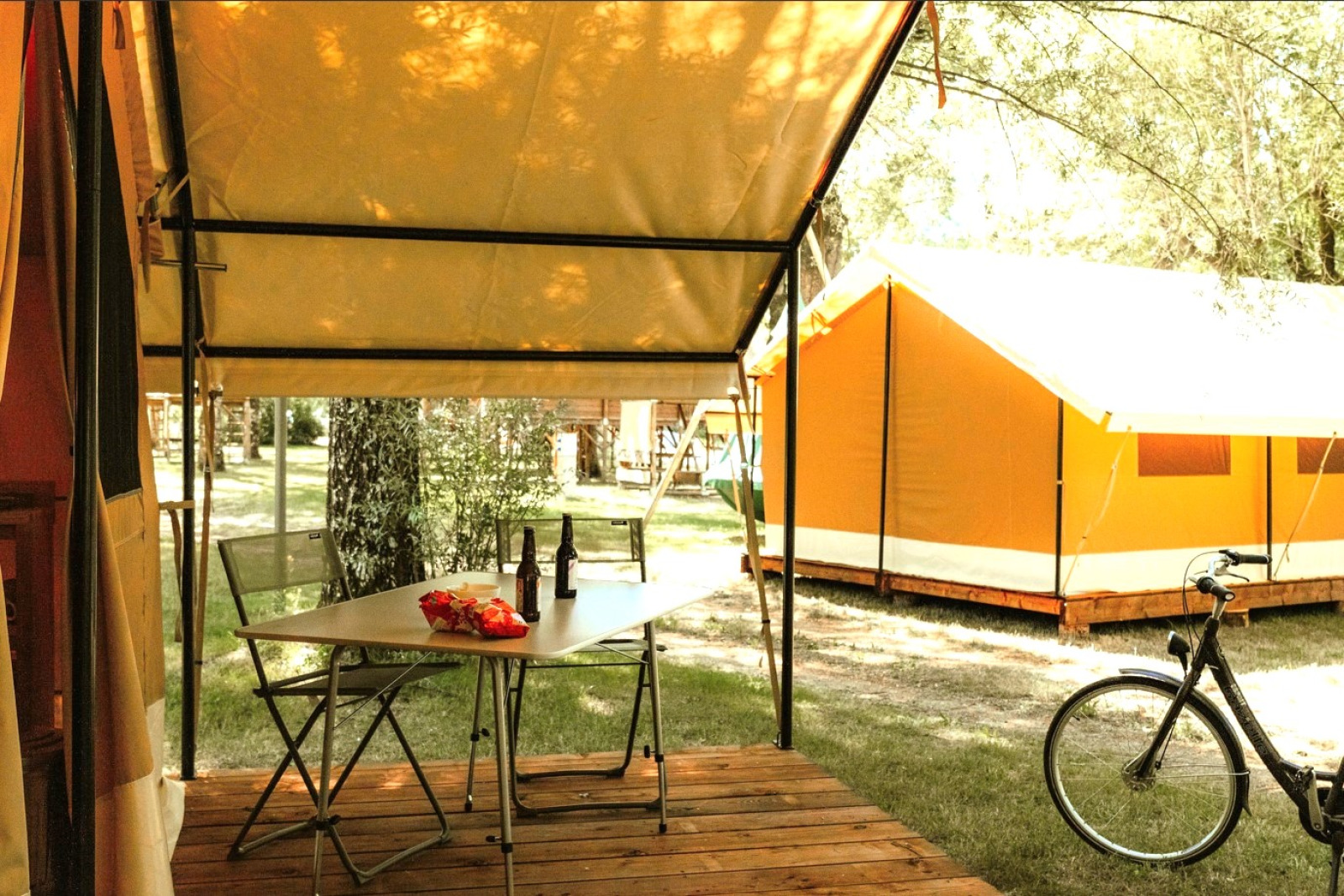Safari-tent met buitenterras, tafel met snacks en drankjes, fiets en andere tenten op de achtergrond.