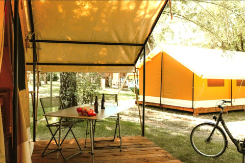 Zona exterior de tienda safari con mesa, sillas, bebidas y snacks, bicicleta y tienda cercana al fondo.