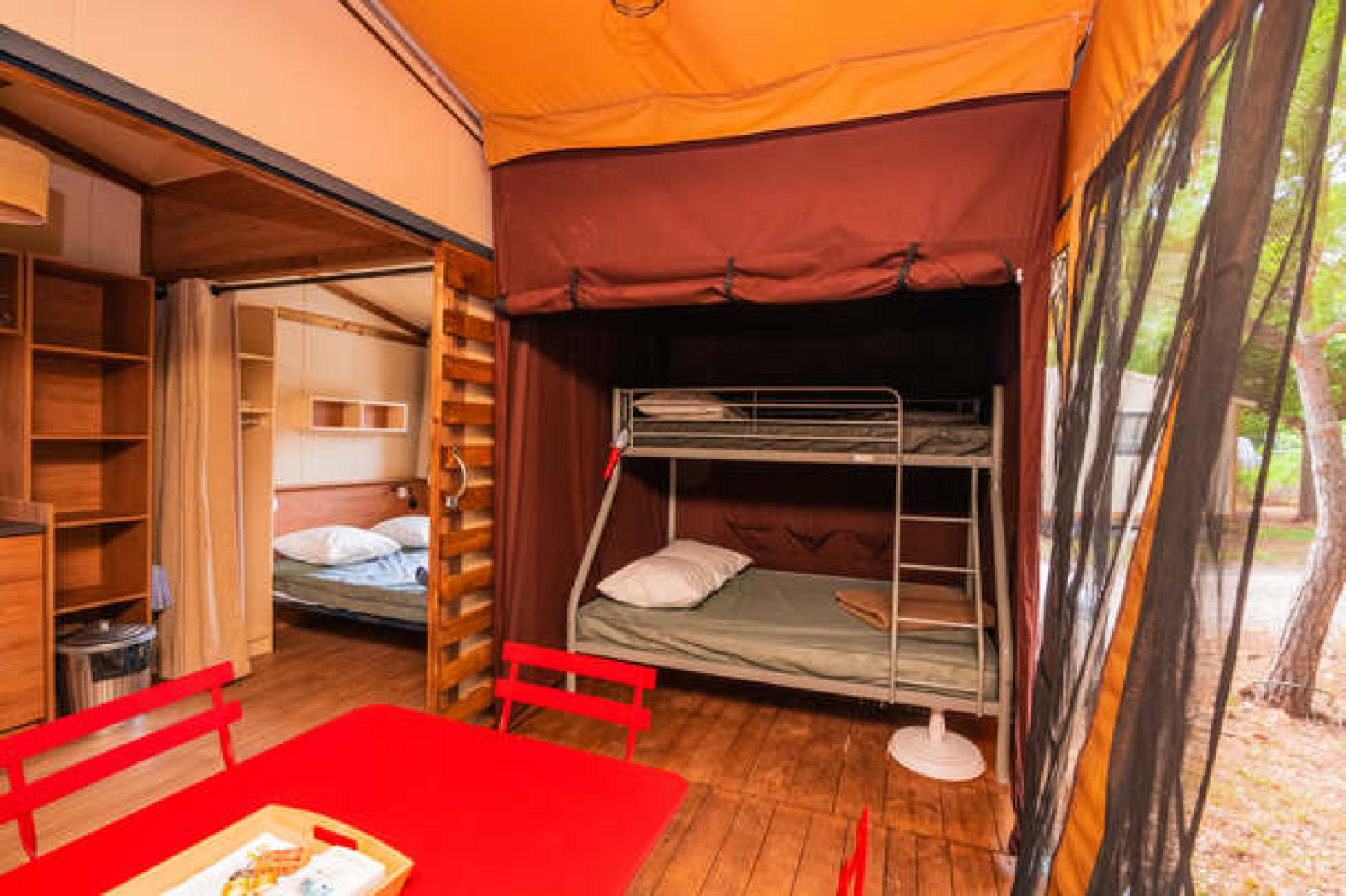 Interieur van een safaritent met stapelbed, tweepersoonsbed en rode eettafel op houten vloer.