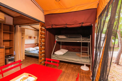 Interior de una tienda safari con literas, cama doble y mesa de comedor roja sobre suelo de madera.