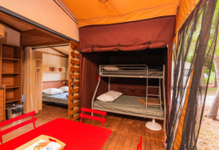 Interior de una tienda safari con literas, cama doble y mesa de comedor roja sobre suelo de madera.