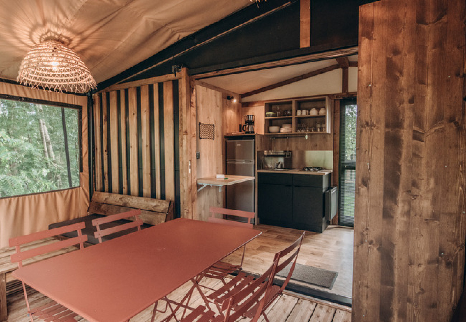 Interieur van een safaritent met eettafel, compacte keuken, houten accenten en zicht op de natuur buiten.